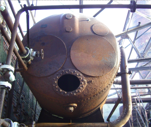 A huge rusty tank, seen from the lower end, looms in an industrial setting of pipes and a smokestack. The end of the tank looks like a face with huge eyes and a black stud-lipped mouth. Definitely scary and looming.